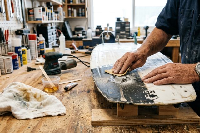 Ponçage d'un impact sur la semelle d'une planche de wakeboard, résine époxy et papier de verre sur un établi