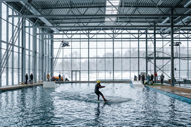 Rider en combinaison sur un câble bi-poulies dans un wakepark couvert, structure métallique et baies vitrées