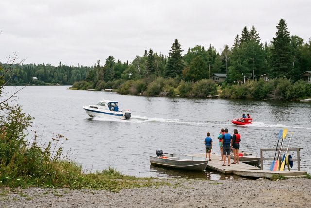 Un bateau tracte une bouée gonflable avec deux personnes sur un lac, vue de loin depuis la berge.